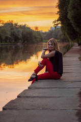 The girl in red trousers sits on the embankment of the river at sunset