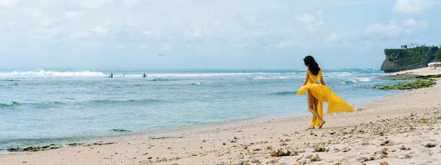 Woman in long yellow dress on rocky beach on Bali, Indonesia. Summer time. Dark hair brunette