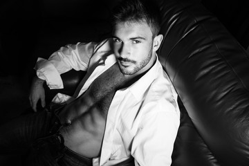 Portrait of attractive young man with beard and open shirt revealing sixpack abs, sitting in...