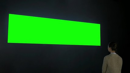 Woman looking at wide large wall blank interactive green display in dark room of modern technology exhibition or museum. Mock up, green screen, futuristic, template, education, chroma key concept