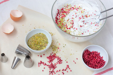 Preparation of meringue with pistachio and raspberry in a glass bowl. White textile towel and waxed paper on background. Flat lay. Day light
