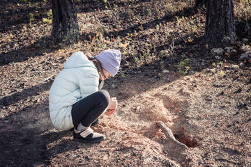 Adult girl in light jacket and hat playing with sand in pine forest.