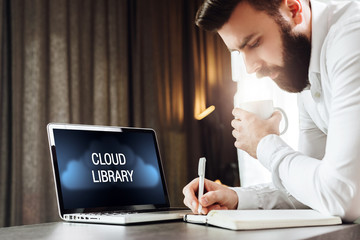 Bearded businessman is standing near laptop with inscription on monitor- cloud library, writing in paper notebook, drinking coffee