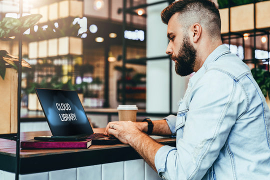 Bearded hipster man sitting in cafe and typing on laptop with inscription on monitor-cloud library. Blogger works in coffee house.