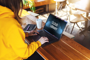 Close-up view of laptop screen with inscription on monitor- cloud library. Girl in yellow hoodie works on computer in cafe.