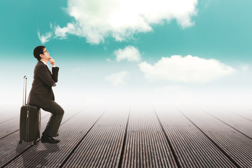 Thoughtful young asia businessman sitting on travel bag holding hand on chin and looking away to white cloud thought bubble
