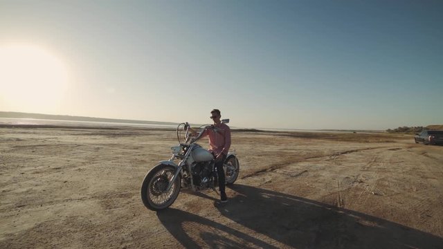 Young stylish motorcyclist goes to his bike in the desert road during sunset 