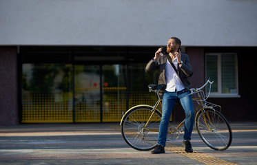 A young stylish bearded  businessman in suit going to work by bike. Drinking coffee from a cup to go and talking on a mobile  phone on the background of an office building. Takes a working break