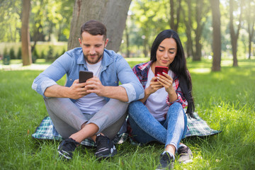 loving couple sitting on a plaid blanket in the park and use the phone, handsome guy and girl