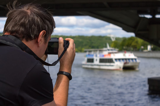 A tourist takes pictures of boats sailing past him along the river under a bridge
