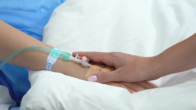 Mother stroking daughter hand with drip lying in hospital bed, family support
