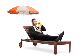 Young businessman in a suit lying on a sunbed under umbrella and sipping a cocktail