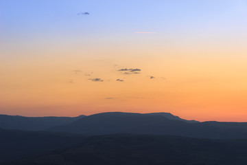 Cold to warm gradient on a sunrise sky above silhouette mountains, small dark clouds and sunlit plane trails