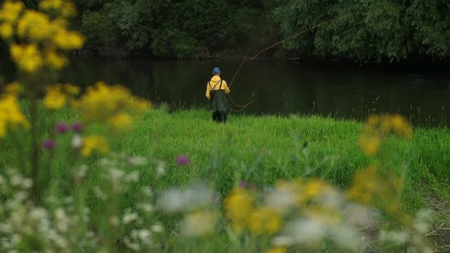 male fisherman in yellow jacket and blue cap special clothes, throws a float, fishing on the river, standing on the shore, on green grass, beautiful nature, in the summer day, Wide angle, slow motion