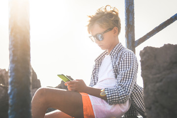 Trendy boy sitting on a stone staircase using smartphone. Serious teen texting with friends online. Backlight portrait of a young male ready to go to the beach. Warm filter. Soft focus on his face.