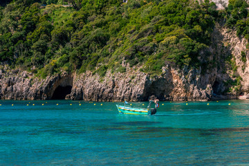 Landscape with turquoise calm sea water, mountain with rocky hillside covered with green trees and bushes and caves, cruise touristic boat. Corfu Island, Greece. 