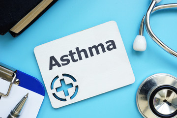Text sign showing Asthma. The text is written on a small wooden board with red cross silhouette. There are pills , stethoscope, blister, blue table on the photo.