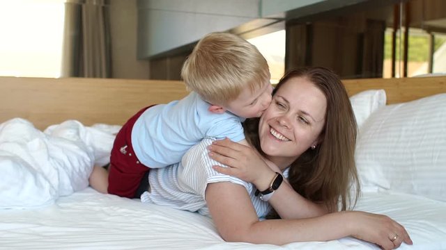 Mom and son lie on the bed in the morning on the weekend and play, the boy kisses and hugs his beloved mother. Slow motion.