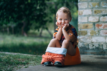 little child sits on a pot in the garden. He is smiling and looking at the frame.