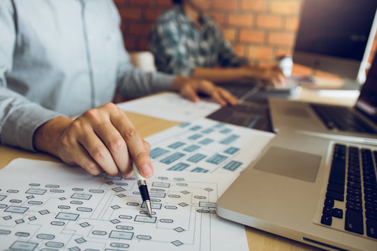 Software developer is pointing to the paper working and analyzing together the code written into the program on the computer.