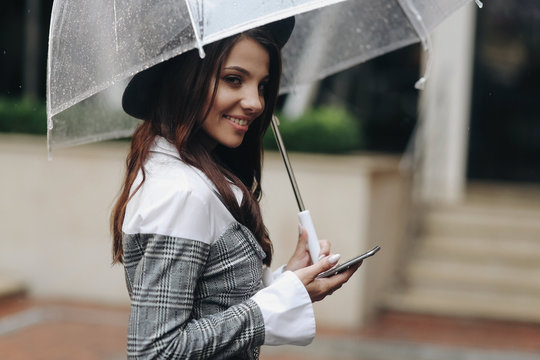 Portrait of beautiful woman wearing black hat and gray coat under transparent umbrella, holding smartphone in your hand