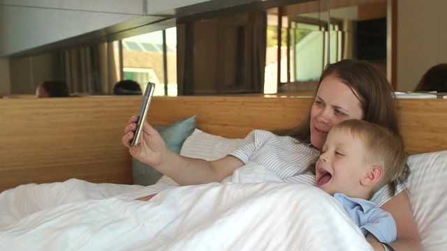 Happy smiling young woman with her child taking a selfie on the phone lying on the bed in the morning. Mother and son smile and take pictures of themselves on the smartphone.