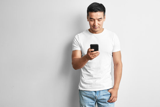 Handsome Asian guy in white studio smiles joyfully looks at the screen of mobile phone. Happy contented man dressed casual writes and reads sms on smartphone
