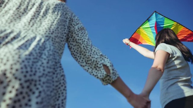 Close-up of two happy young caucasian women holding hands staying outdoors. Low angle view pretty bisexual girls flying a colorful kite in blue skyline. Love freedom. LGBT.