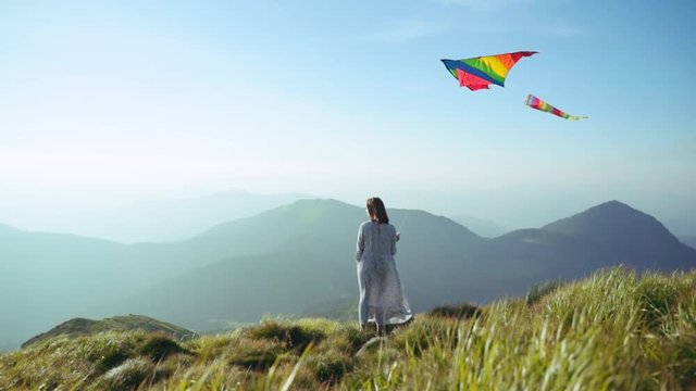 joyful young woman in dress holding rainbow kite standing on mountain peak. view of happy girl traveler enjoying summer nature and sunlight in highland.