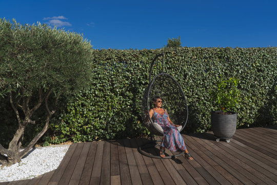 a woman sit in a suspended seat on a wooden floor in a garden with an olive tree