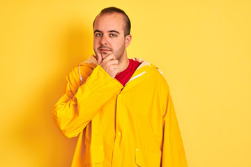 Young man wearing rain coat standing over isolated yellow background looking confident at the camera with smile with crossed arms and hand raised on chin. Thinking positive.