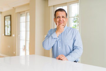 Fototapeta premium Middle age man sitting at home looking confident at the camera with smile with crossed arms and hand raised on chin. Thinking positive.