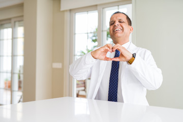 Fototapeta premium Middle age doctor man wearing medical coat at the clinic smiling in love showing heart symbol and shape with hands. Romantic concept.