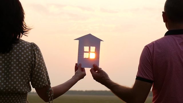 The silhouette of a paper house in the setting sun. A man and a woman holding a paper house and watching the sunset. A happy family dreams.