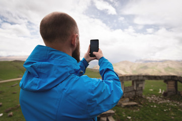 Fototapeta premium A bearded man takes photos or videos outdoors on his smartphone. A guy in a jacket on a background of mountain valleys and a gray sky weedt broadcast on the phone. Back view