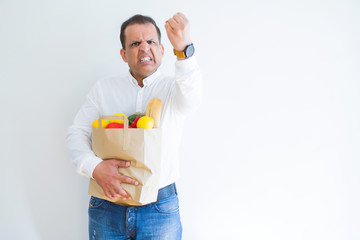 Middle age man holding groceries shopping bag over white background annoyed and frustrated shouting with anger, crazy and yelling with raised hand, anger concept