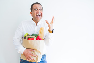 Middle age man holding groceries shopping bag over white background very happy and excited, winner expression celebrating victory screaming with big smile and raised hands