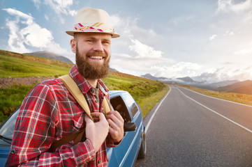 Fototapeta premium Portrait of a bearded happy smiling traveler hipster with a backpack in a plaid shirt and a hat next to an unknown car stands on the road at sunset in the mountains. Happy and confident travel concept