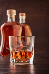 A glass of whisky with ice on a wooden table. In the background, two bottles of whiskey of different shapes.