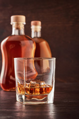 A glass of whisky with ice on a wooden table. In the background, two bottles of whiskey of different shapes.