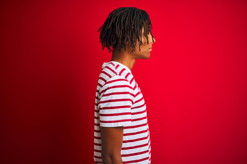 Young afro man with dreadlocks wearing striped t-shirt standing over isolated red background looking to side, relax profile pose with natural face and confident smile.