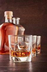 Three glasses of whiskey on the ice on the wooden table. In the background are two bottles of whiskey, full and open.