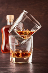 Two glasses of whisky with ice on a wooden table. One glass stands on the other. There's a full bottle of whiskey in the background.