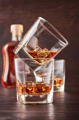 Two glasses of whisky with ice on a wooden table. One glass stands on the other. In the background is a full bottle of whiskey and a glass of whiskey.
