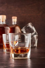 Two glasses of whisky with ice on a wooden table. In the background there are two bottles of whiskey of different shapes and empty glasses for whiskey.