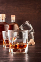Two glasses of whisky with ice on a wooden table. In the background there are two bottles of whiskey of different shapes, empty glasses for whiskey and a wooden stopper for the bottle.