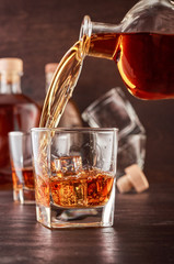 Glass of whiskey on a wooden table, which poured whiskey from a bottle. In the background, two bottles of different shapes, full and empty glasses of whiskey.