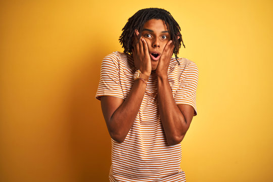Afro man with dreadlocks wearing striped t-shirt standing over isolated yellow background afraid and shocked, surprise and amazed expression with hands on face