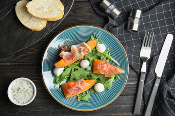 Flat lay composition of fresh melon with prosciutto, mozzarella and arugula on black wooden table