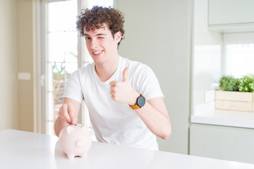 Fototapeta na wymiar Young man investing money using piggy bank at home happy with big smile doing ok sign, thumb up with fingers, excellent sign